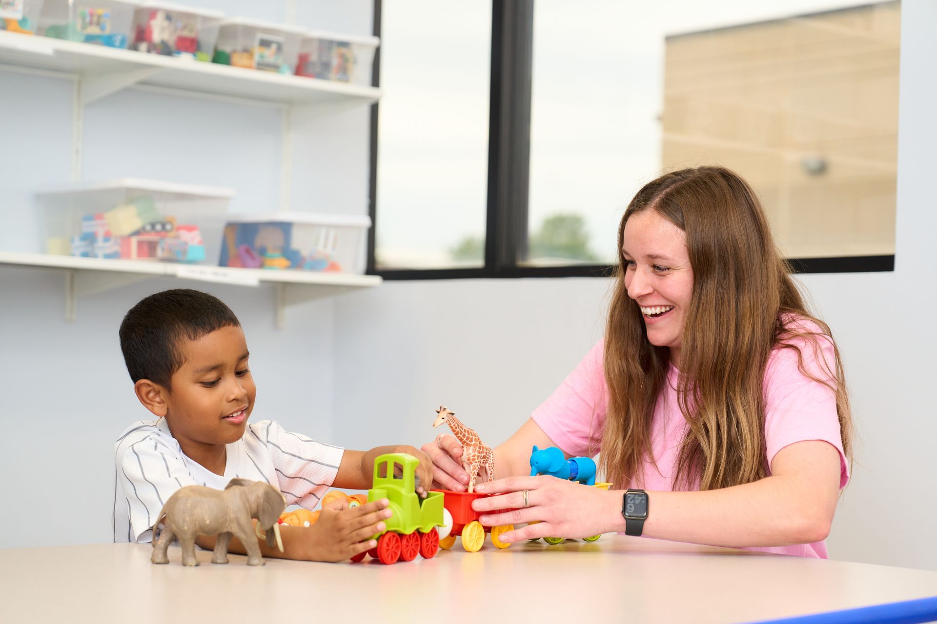 Two children playing with colorful toys at a table in a bright classroom, smiling and interacting.