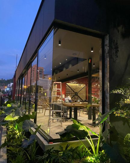 Terraza exterior de un restaurante al atardecer, con luces cálidas, paredes de cristal y exuberante vegetación.