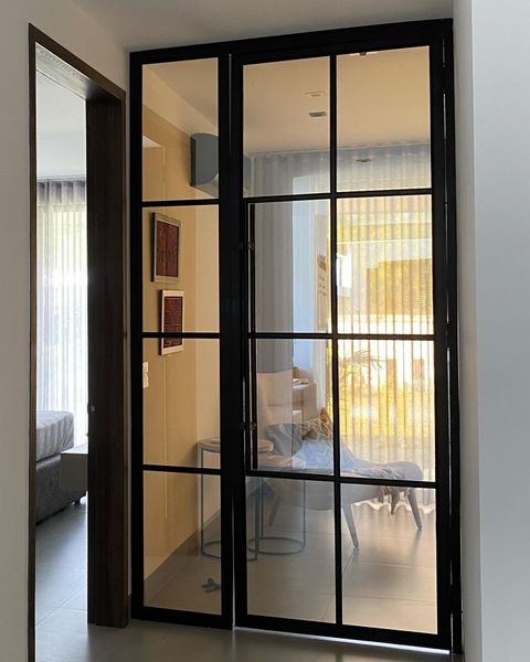 Pasillo estrecho con puerta de cristal con marco negro que da a una habitación luminosa con sofá y silla.