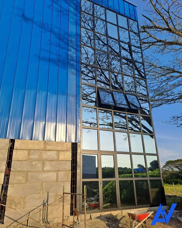 Fachada de edificio moderno con paneles de vidrio azul y ventanas reflectantes junto a un muro de piedra y árboles.