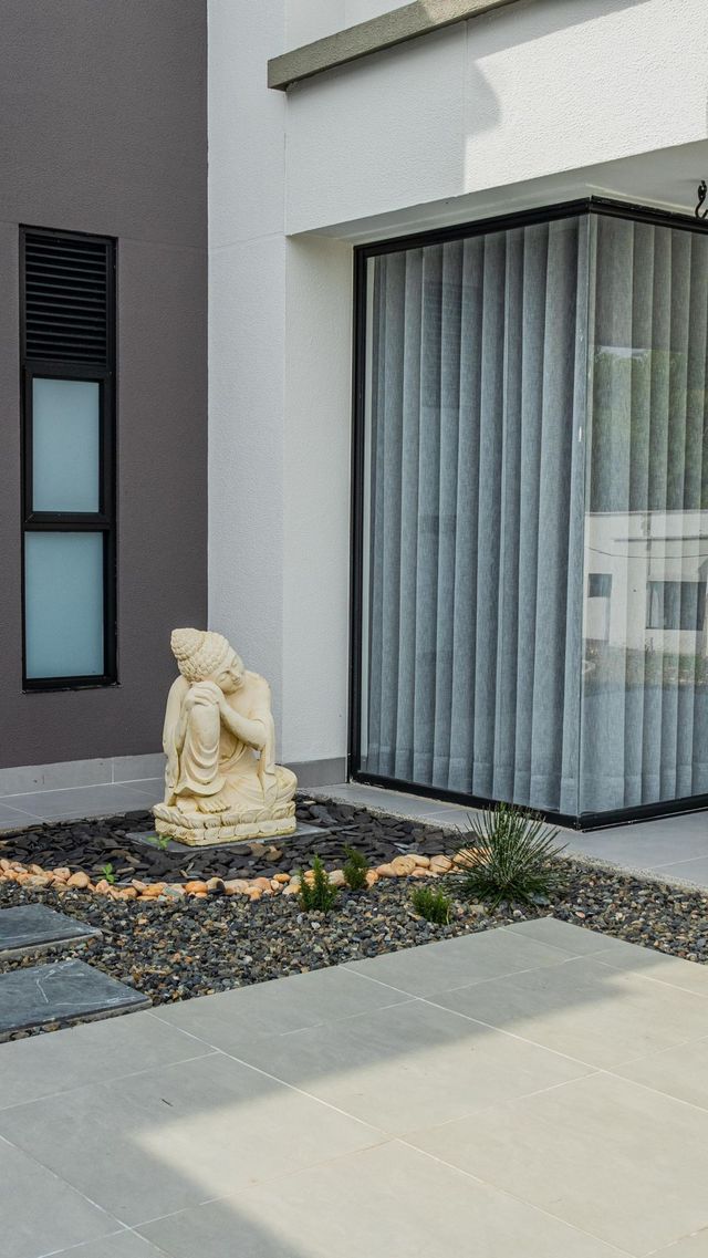 Una pequeña estatua de jardín junto a la entrada de una casa moderna con grandes ventanales y un camino pavimentado.
