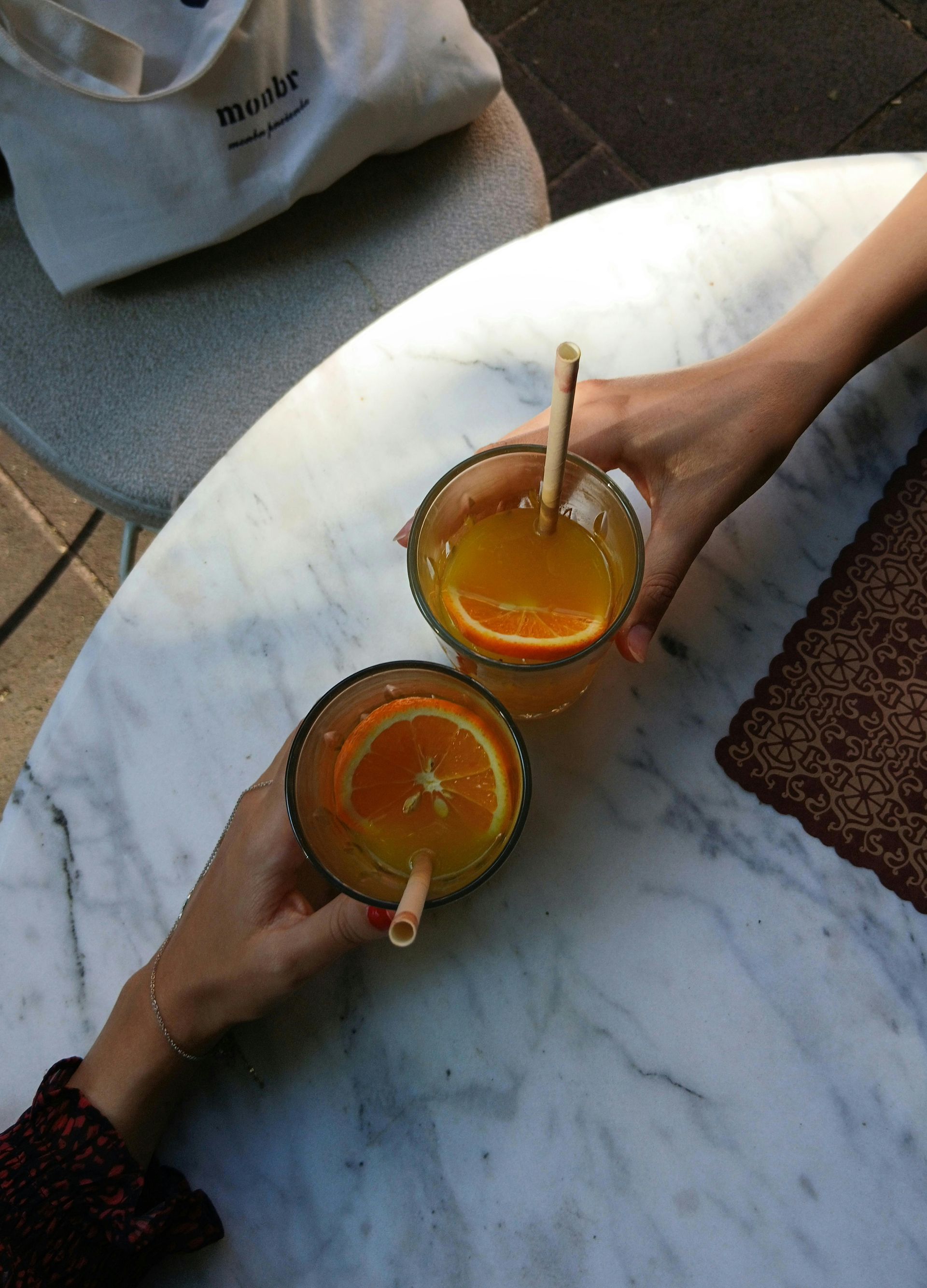Two hands holding glasses of orange drinks on a marble table, with orange slices and straws.