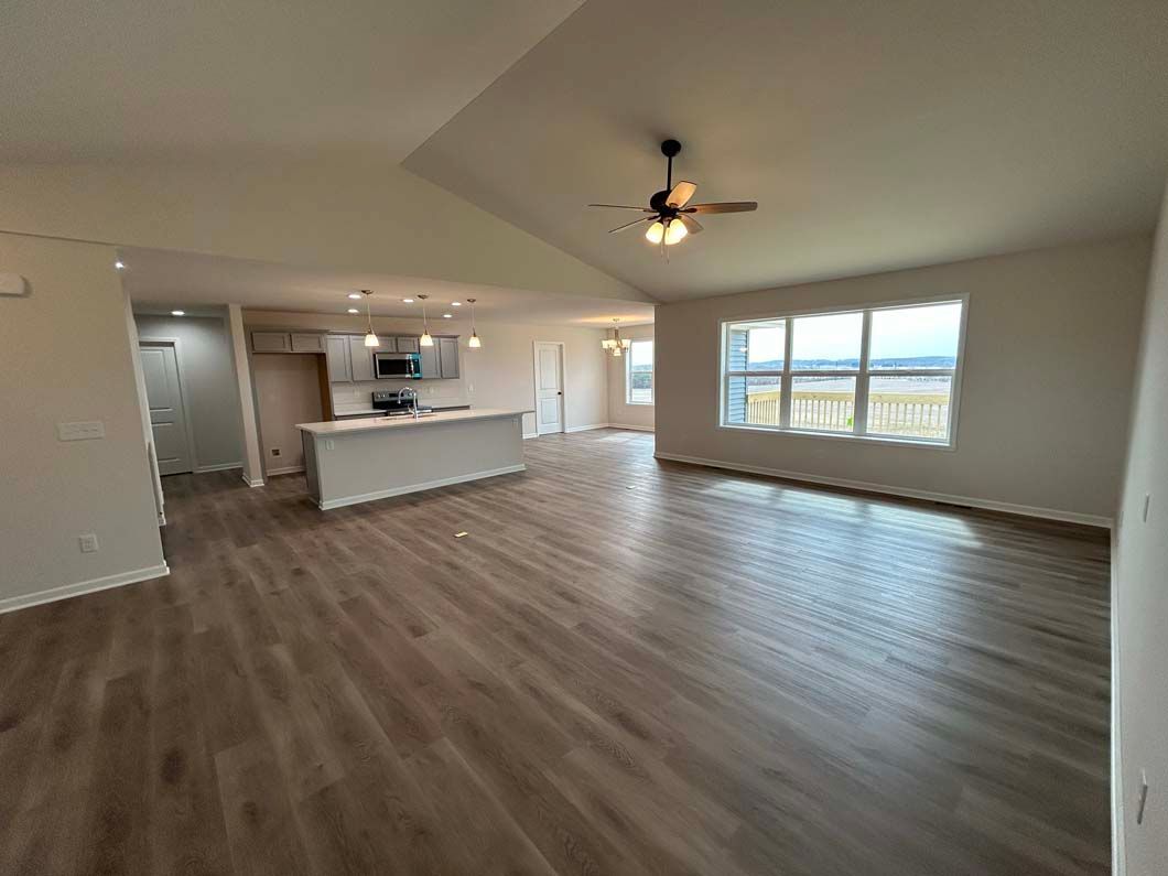 Spacious, empty living room with wooden floors, open to a kitchen with an island and window views.