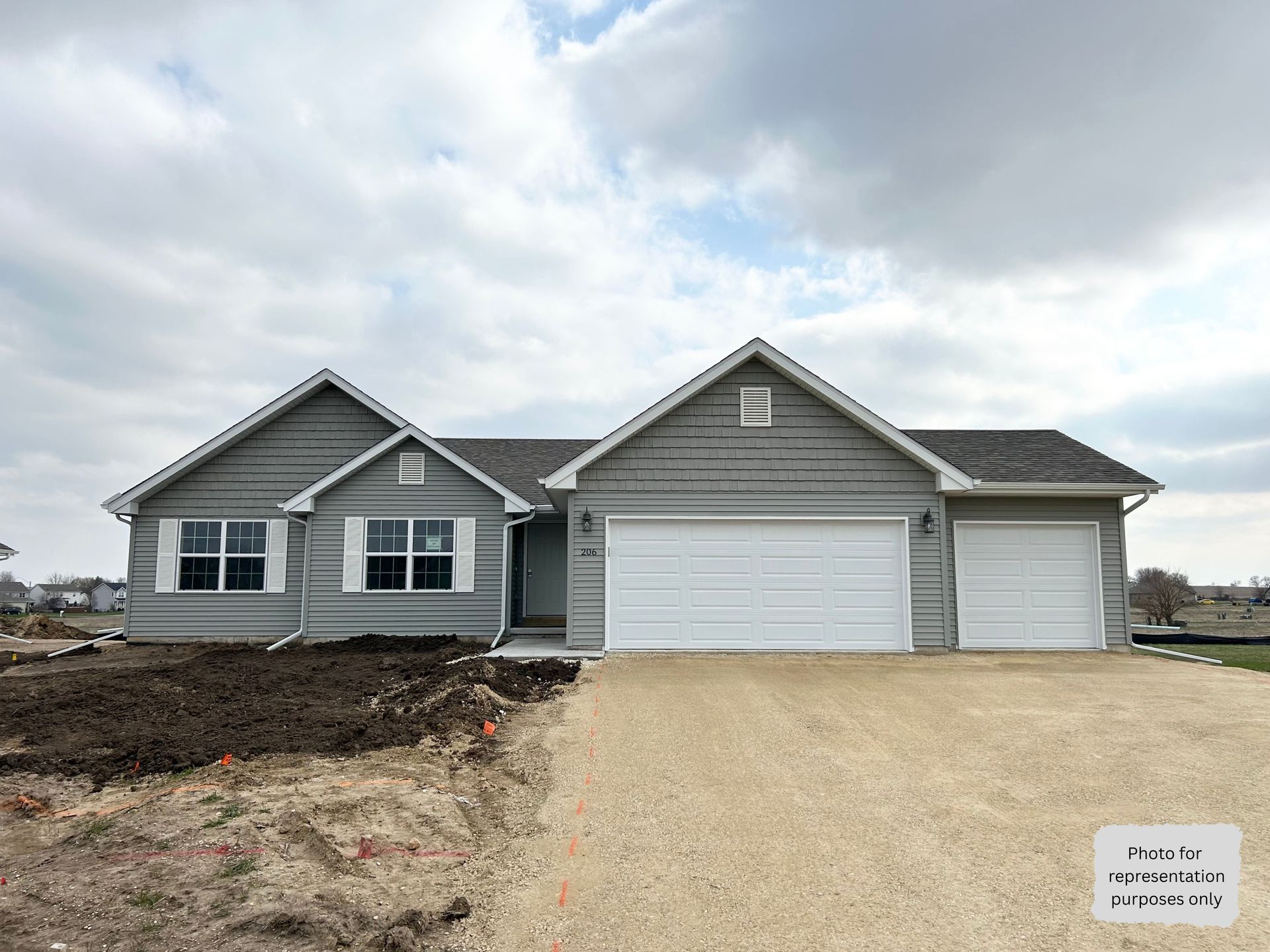 Gray ranch-style house with white garage doors and shutters under cloudy sky; construction site.
