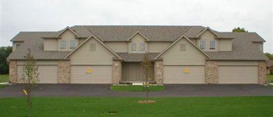 Multi-unit residential building with attached garages; tan siding, asphalt driveway, green lawn.