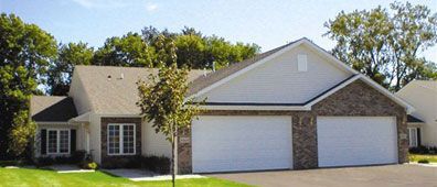 House with white garage doors, brick accents, and green lawn, under a sunny sky.