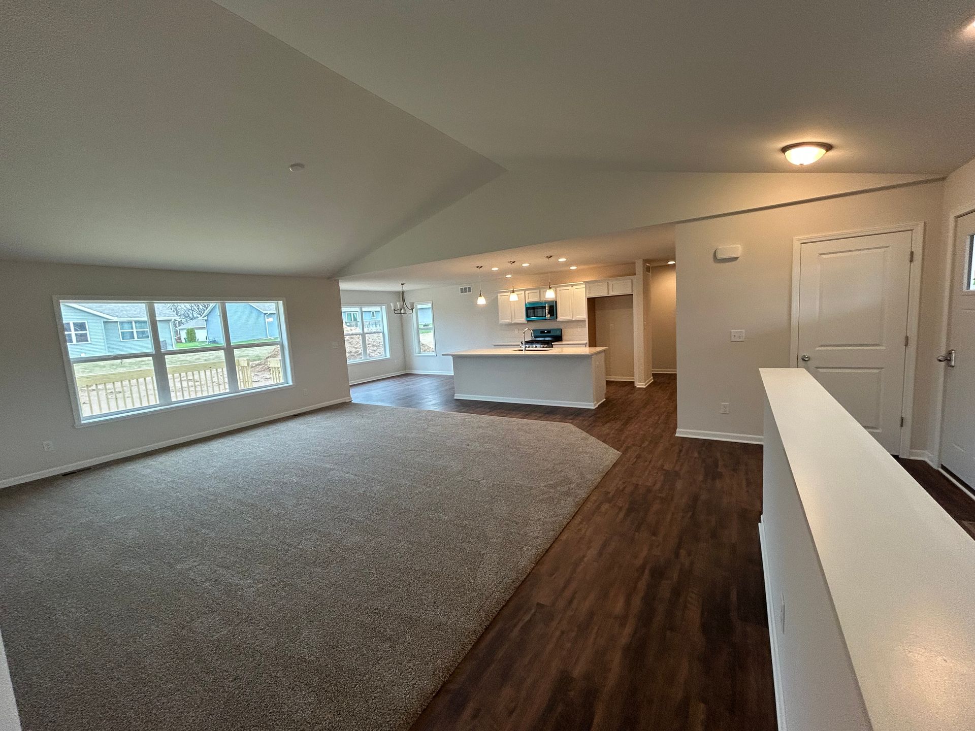 Open-concept living space with neutral tones. Living area with gray carpet, kitchen with white island and dark wood floors.
