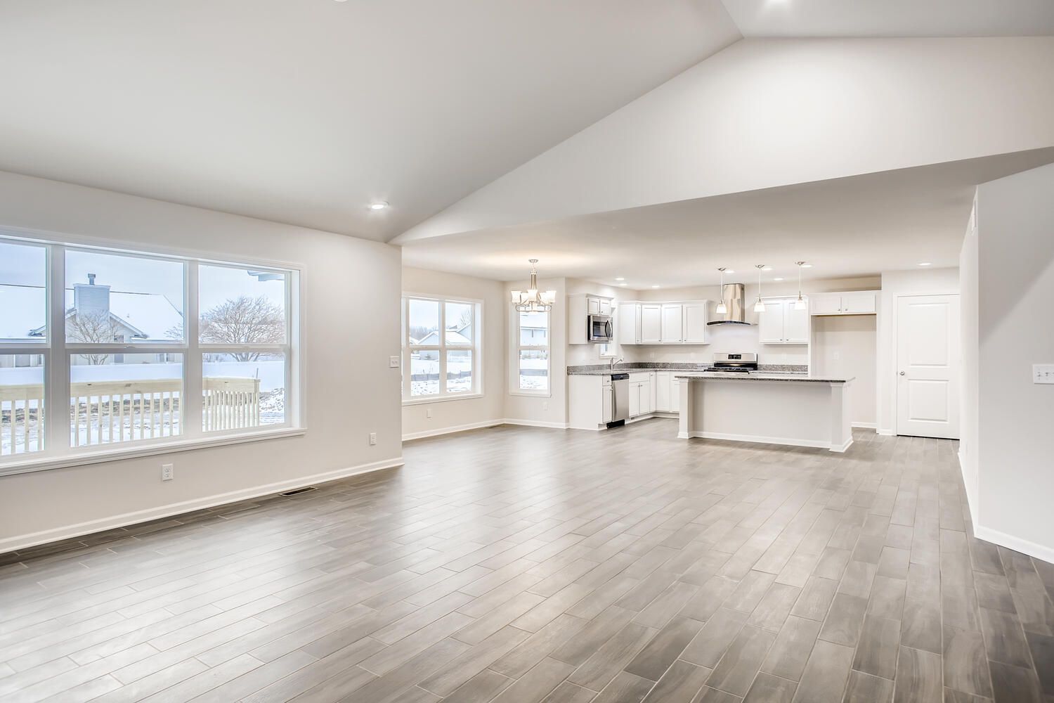 Empty, bright living space with a kitchen in the background, large windows and hardwood floors.