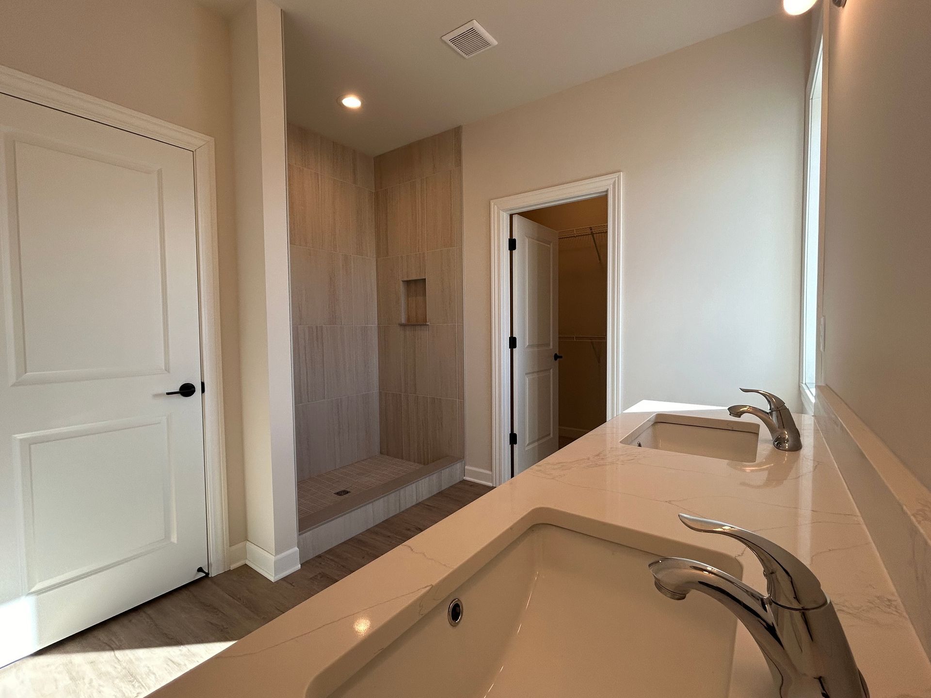 Bathroom with double sinks, shower, and a closed door. Light-colored walls, and a neutral color palette.