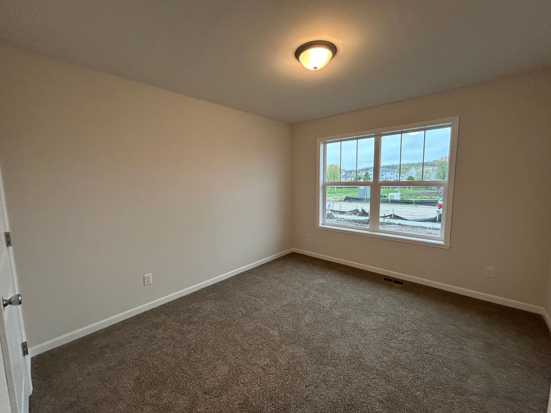 Empty bedroom with beige walls, brown carpet, and a window overlooking a construction site.