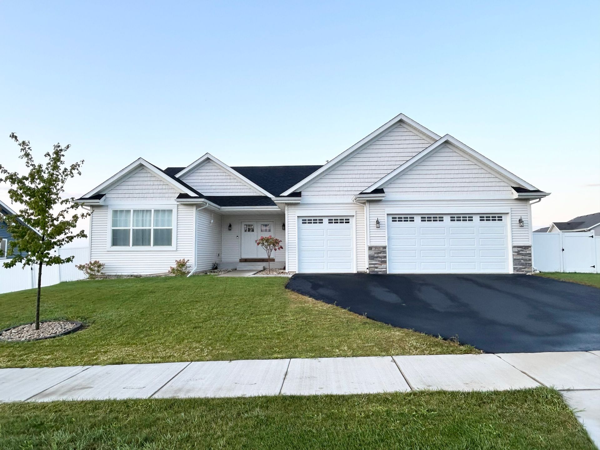 White ranch-style house with a black driveway, green lawn, and a tree. Clear sky.