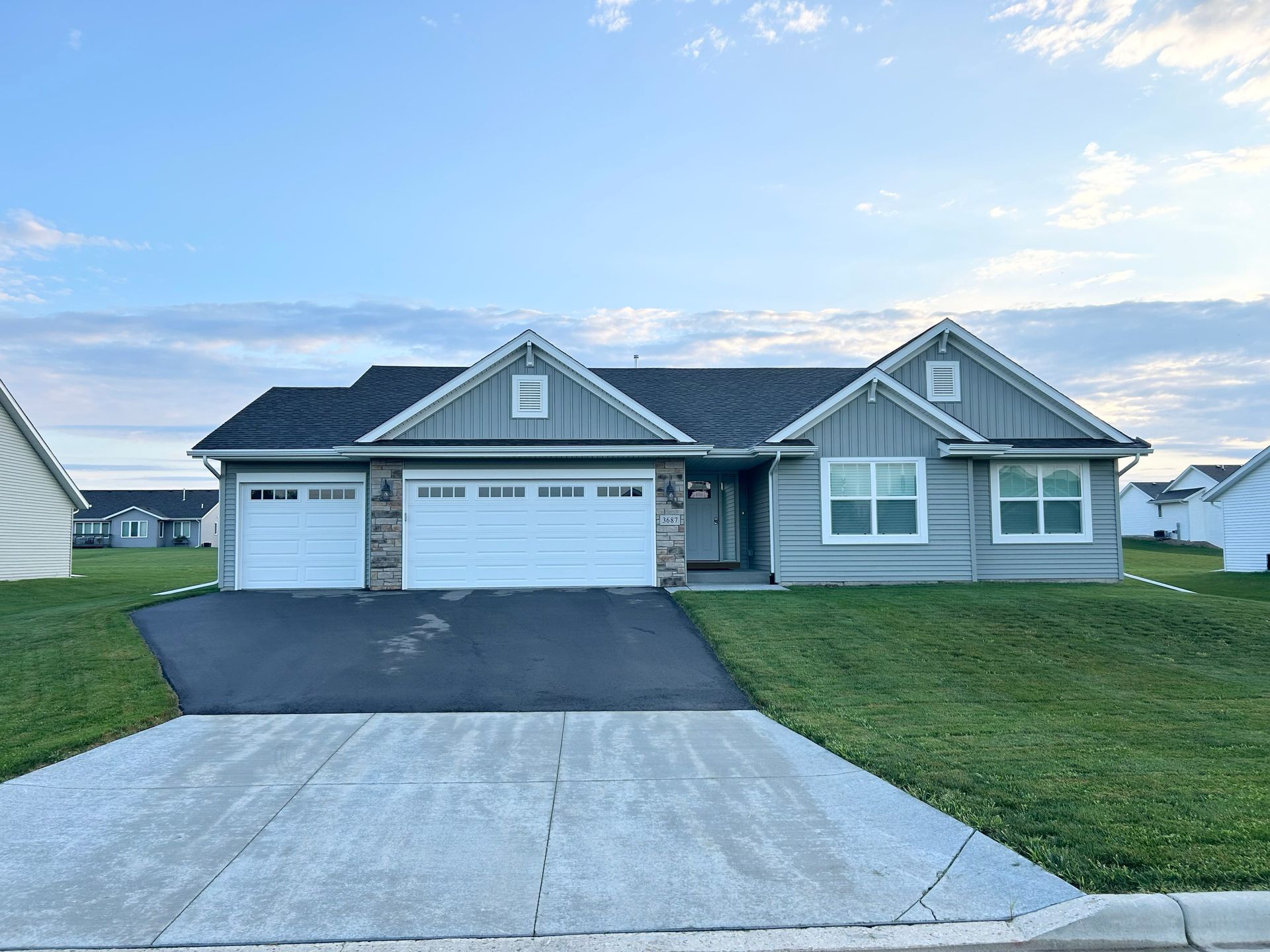 Light blue house with white garage doors, on a grassy hill. Black asphalt driveway, blue sky.