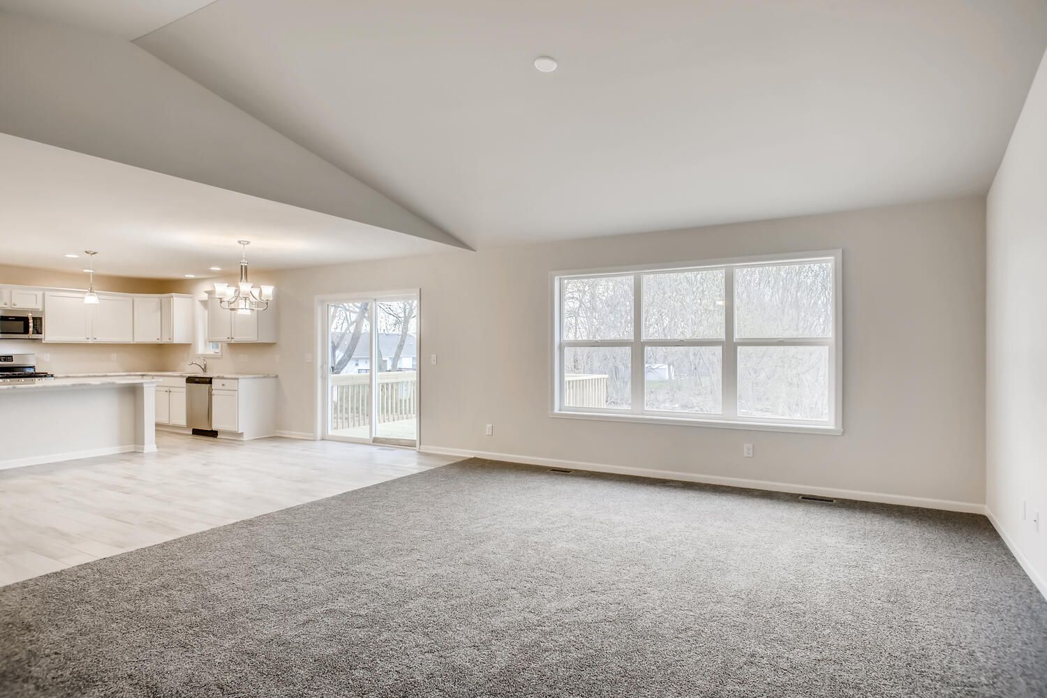 Empty living room with grey carpet, white walls, and a view into a kitchen.