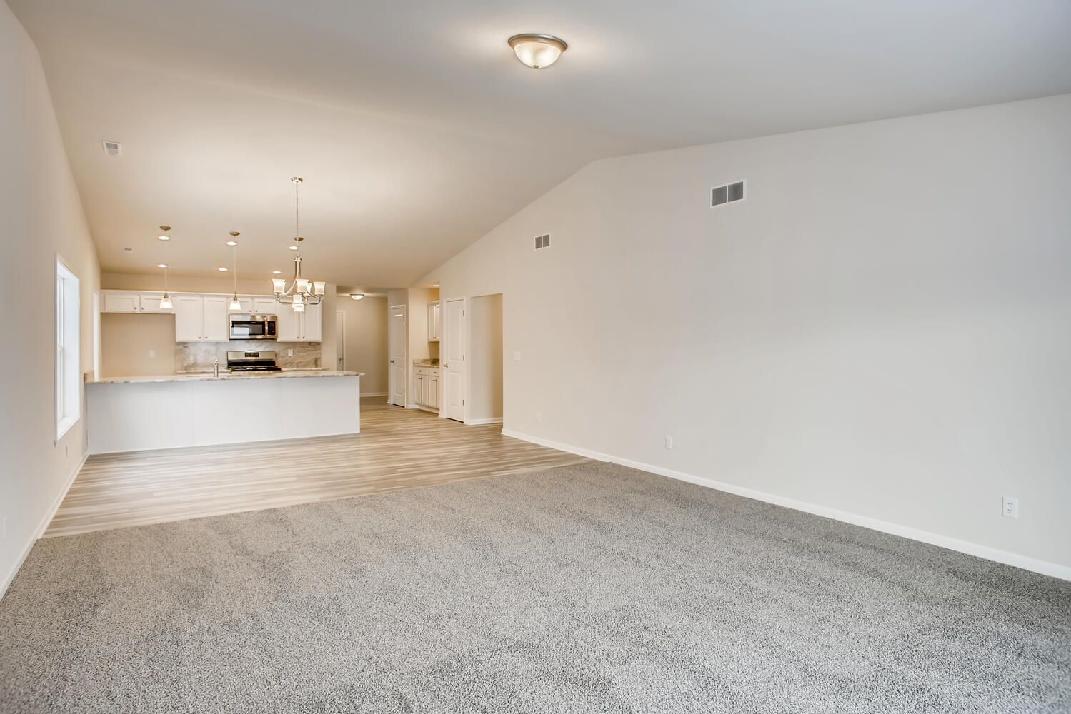 Empty open-concept living space with white walls, light carpet, and kitchen visible in the background.