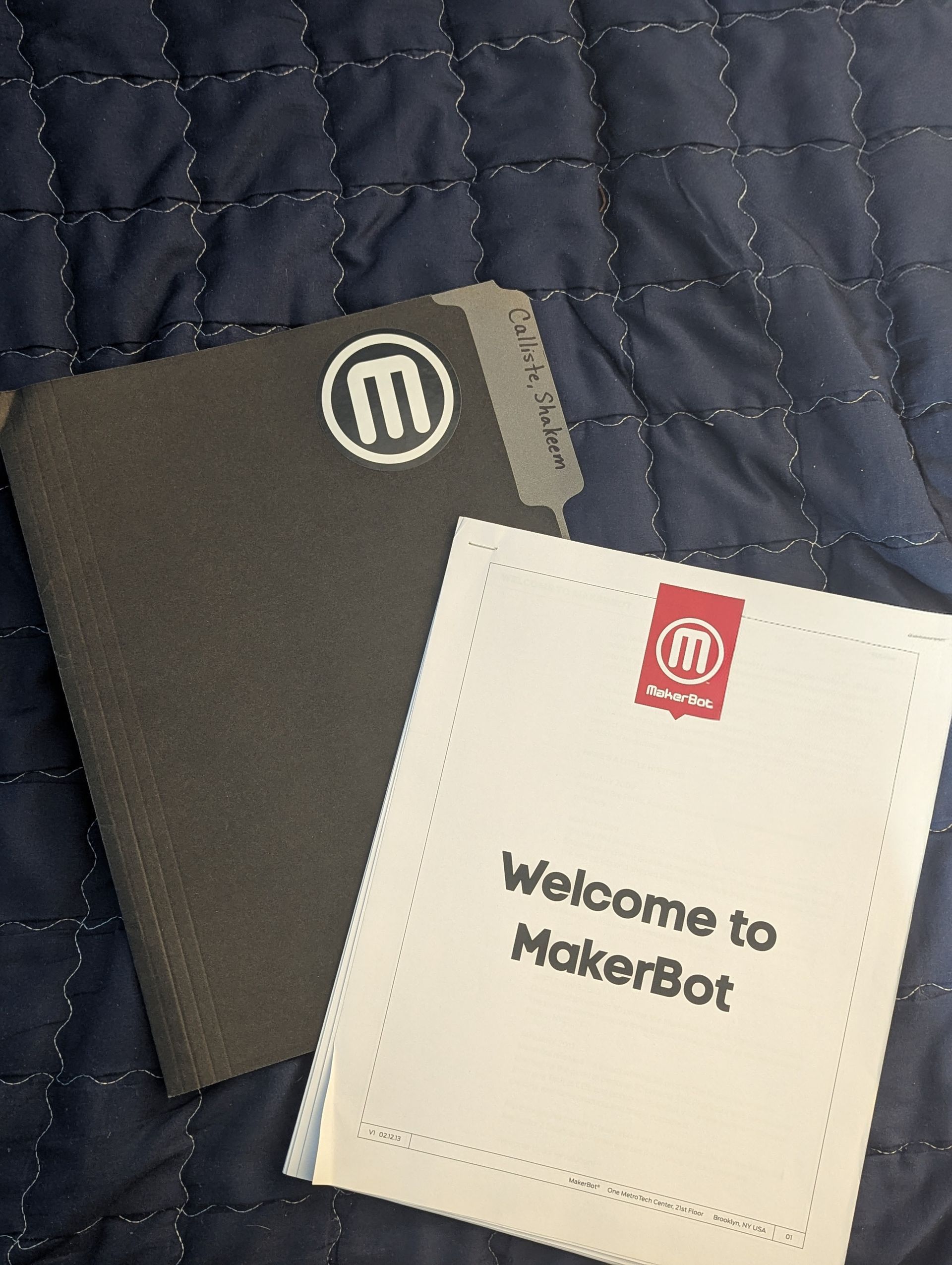 Two MakerBot booklets on a quilted dark blue surface, one black and one white with “Welcome to MakerBot” text.