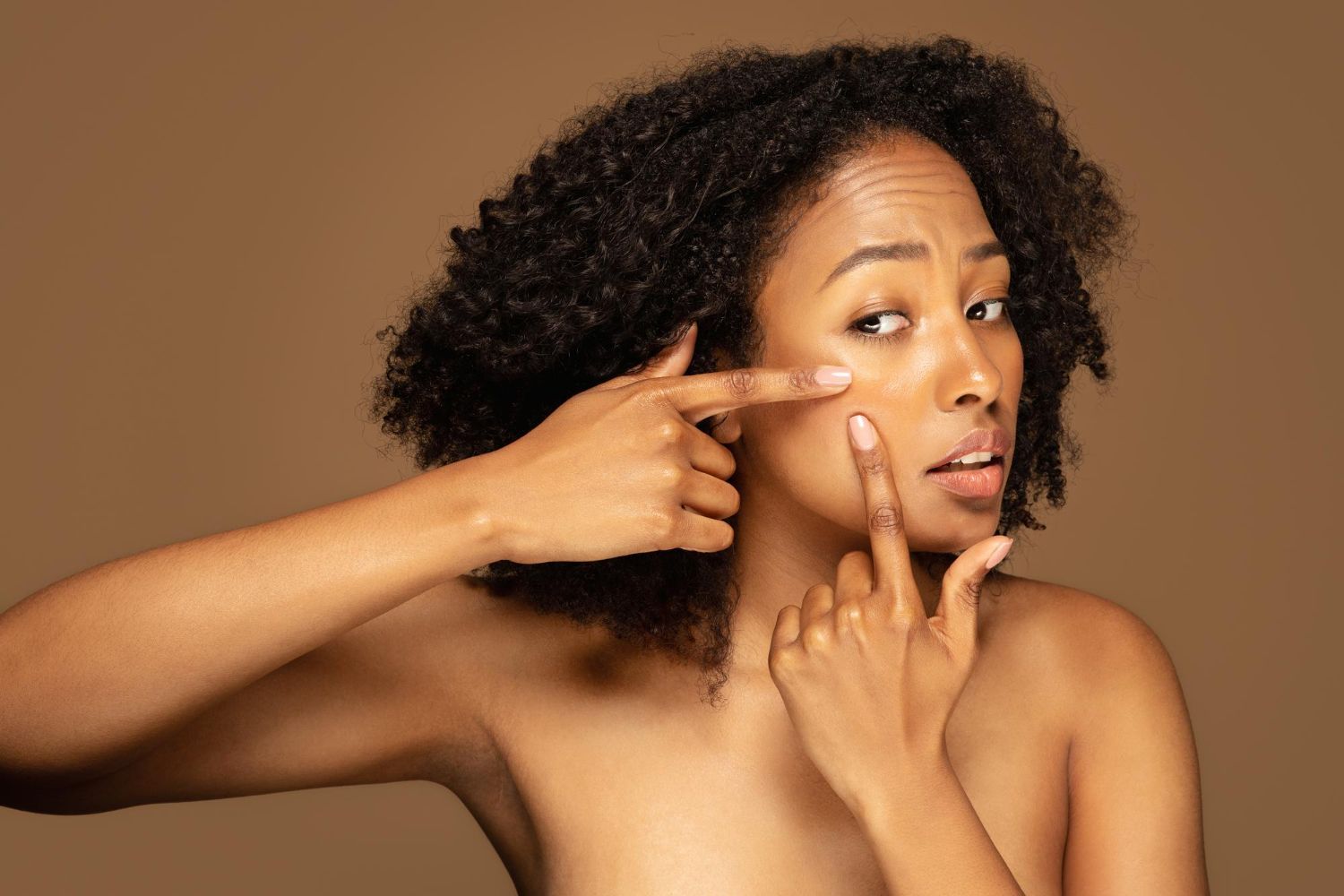 Woman on brown background touching under-eye area and pointing to cheek, showing bare skin near nose and eye