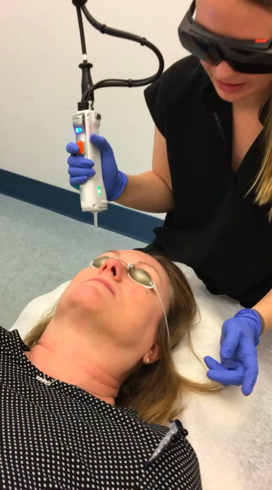 Person in blue gloves uses a device on a woman lying on a treatment table in a clinical setting