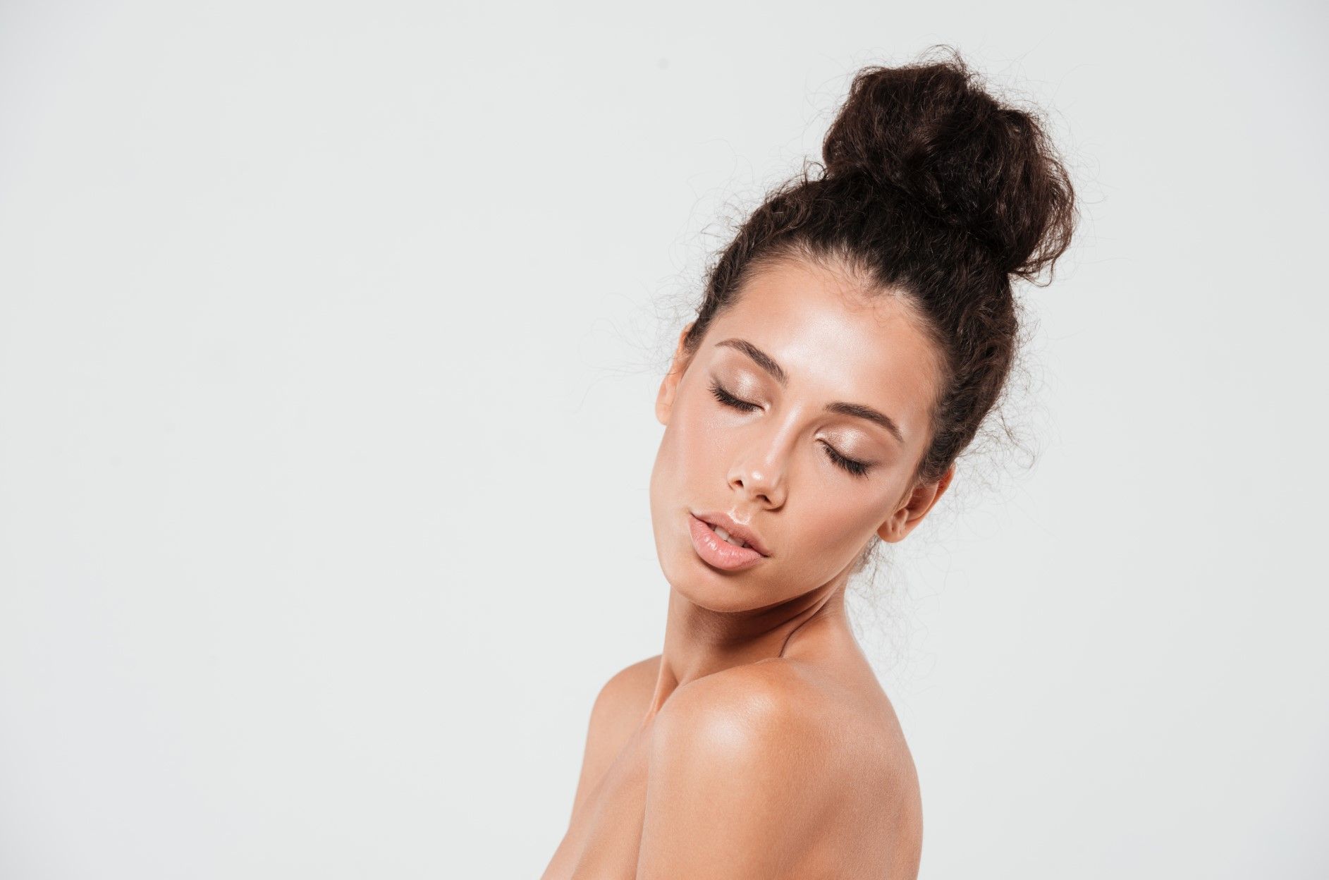 Portrait of a woman with curly hair in a bun, eyes closed against a light gray background