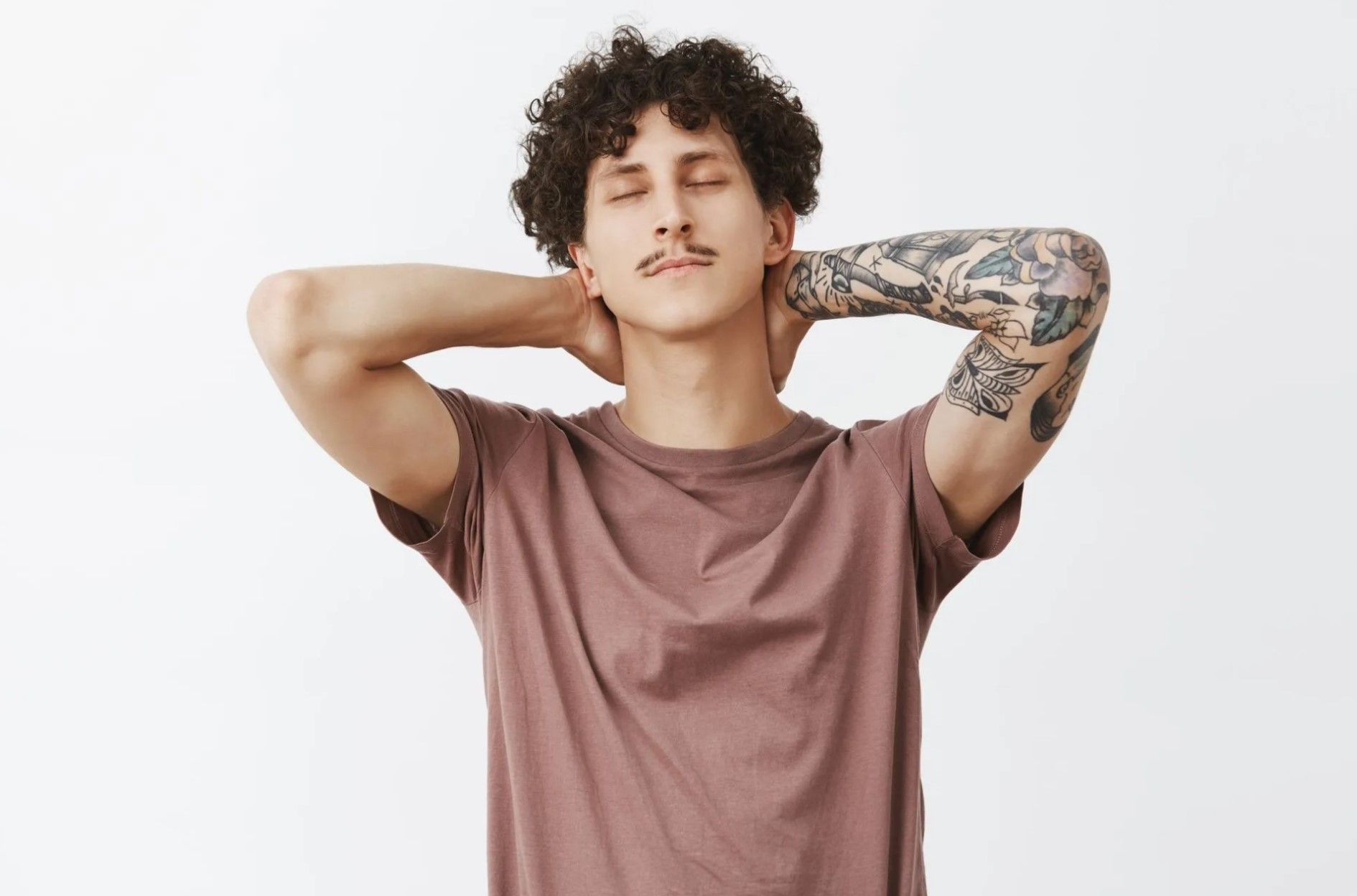 Person with curly hair and tattooed arm stretching with hands behind head against a white background