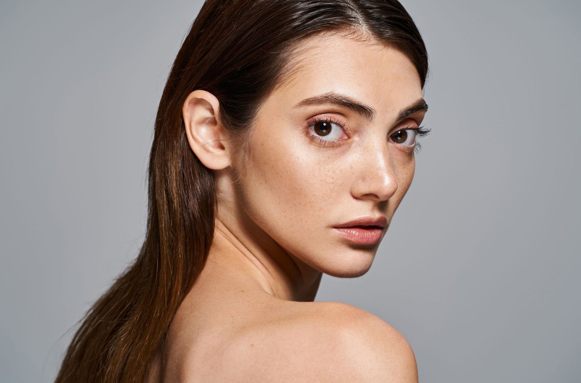 Portrait of a person with long dark hair looking over their shoulder against a gray background