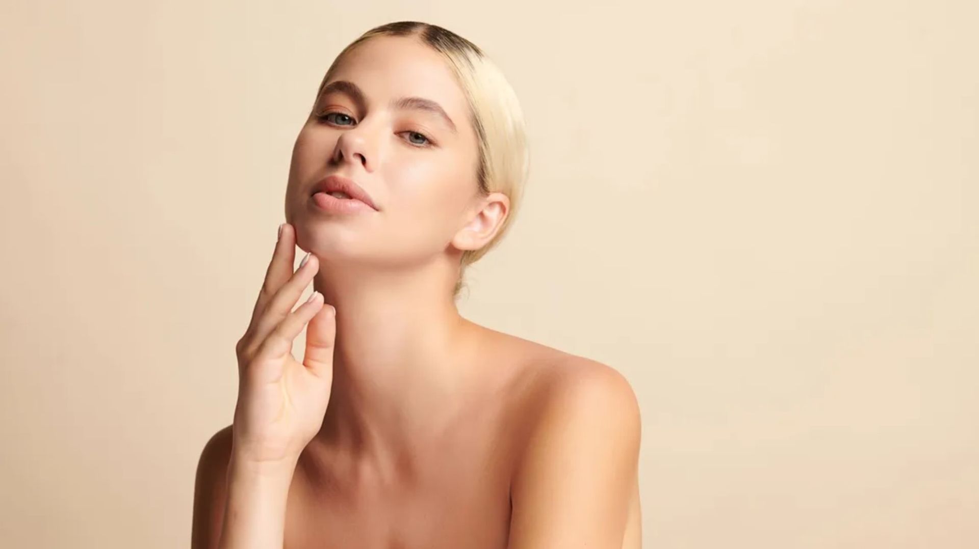 Portrait of a blonde woman with bare shoulders, touching her chin against a beige background