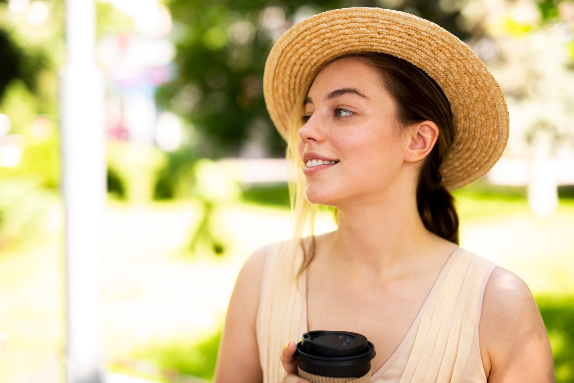 Woman in a straw hat holding a coffee cup outdoors, smiling and looking to the side.