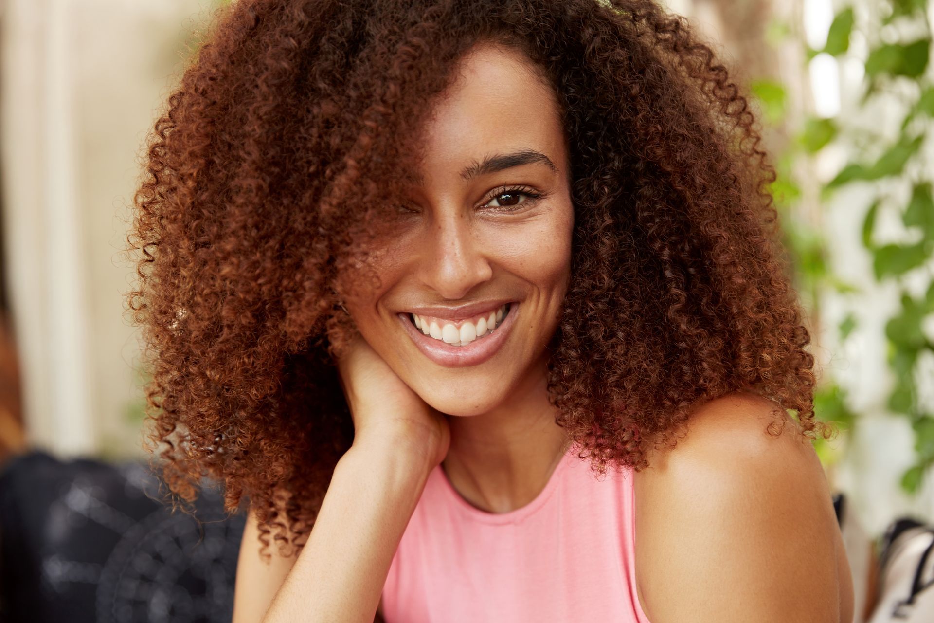 Smiling woman with curly hair in a pink top, resting her chin on her hand indoors