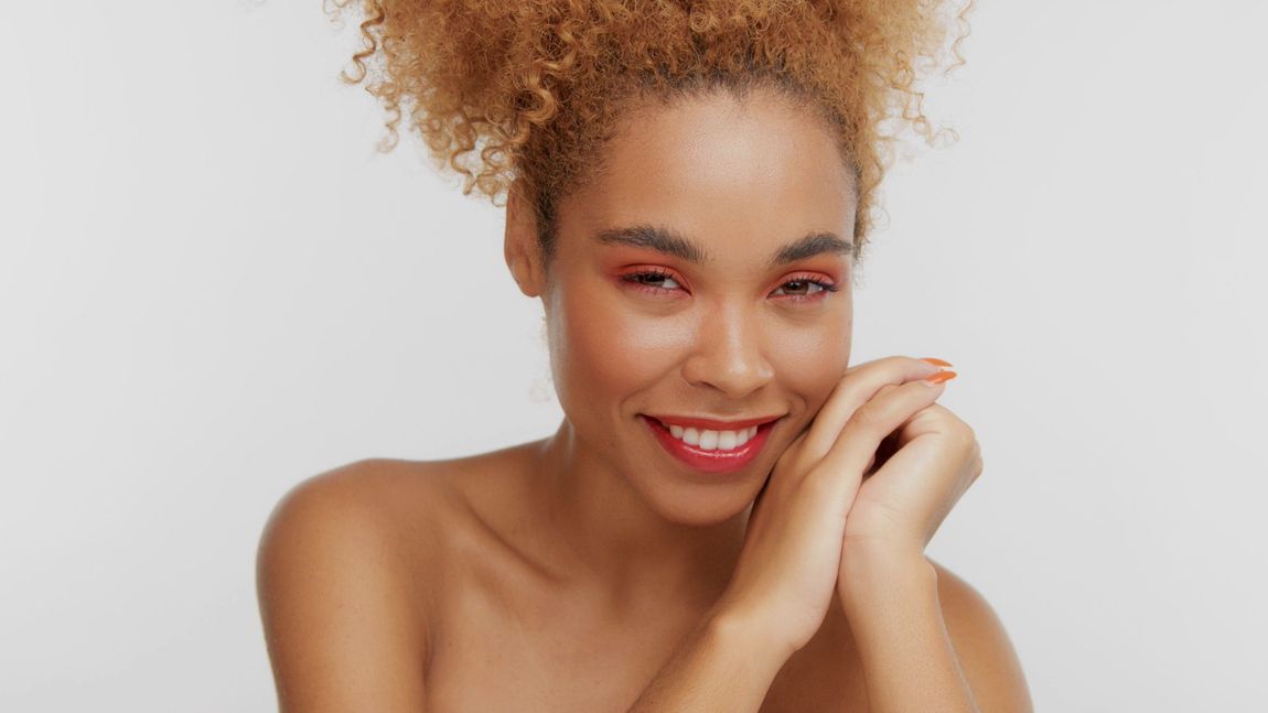 Smiling woman with curly hair resting her chin on her hands against a light background