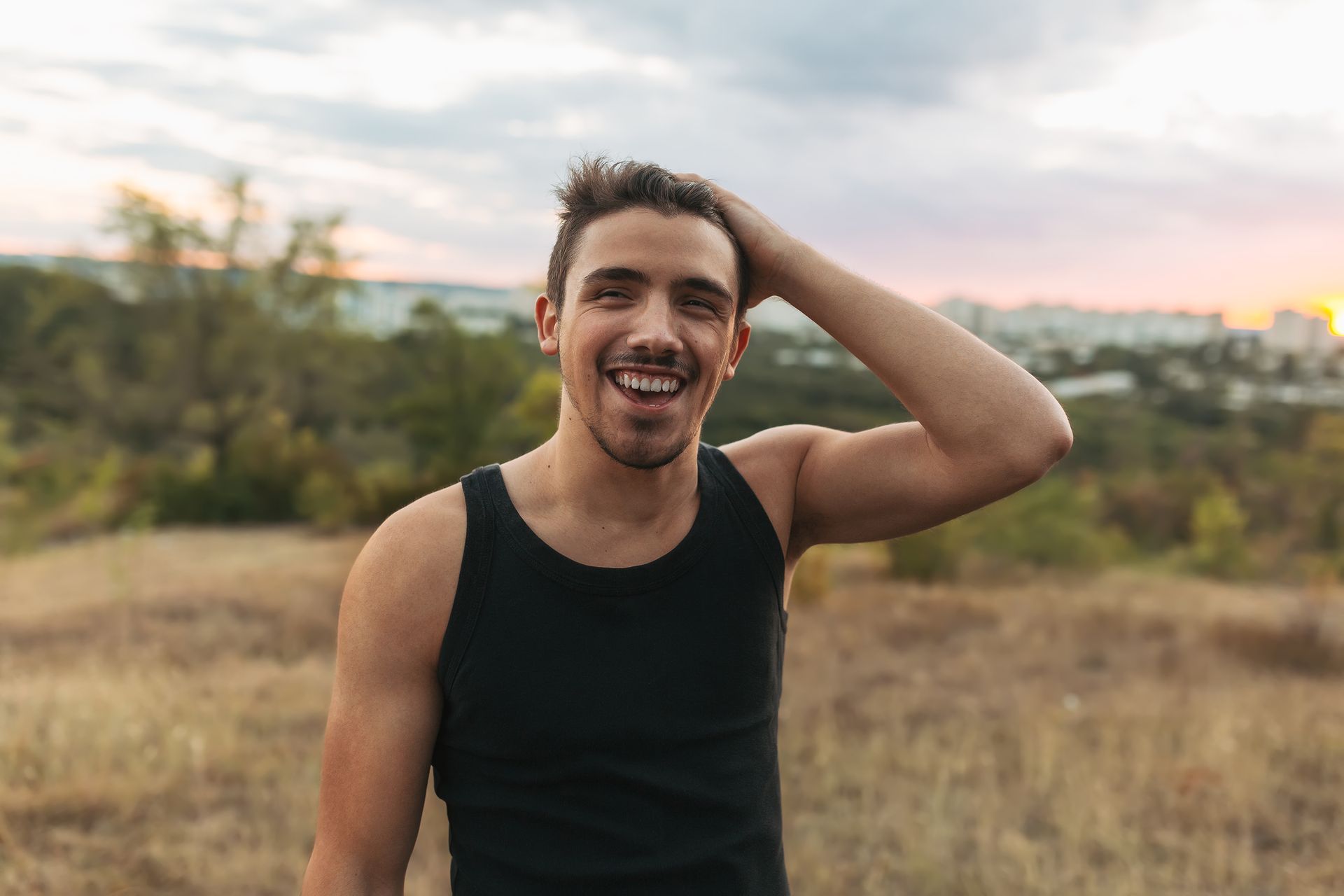 Smiling person in a black tank top standing outdoors at sunset with one hand behind their head