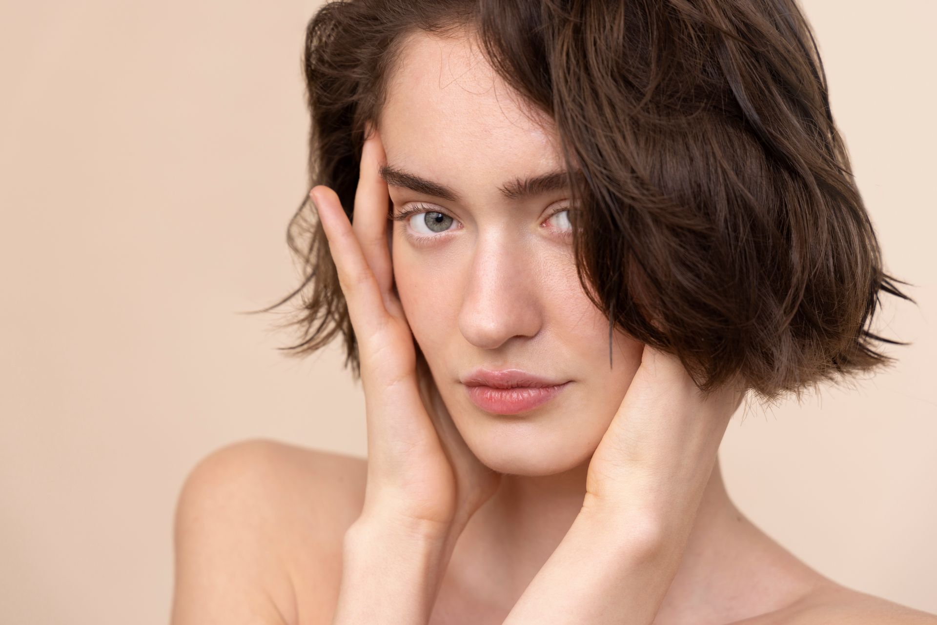 Portrait of a person with short dark hair touching their face against a soft beige background