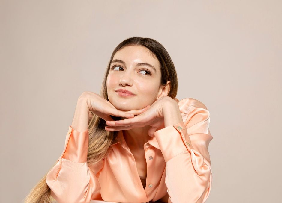 Portrait of a woman in a peach blouse resting her chin on her hands and looking upward with a thoughtful smile