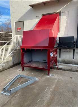 Red trash compactor outside a building with a metal chute, a door, and a nearby gray metal frame.
