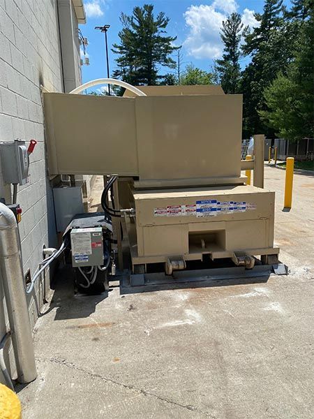 Tan industrial trash compactor outside, next to a building, on a concrete surface.