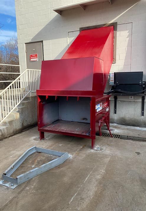 Red industrial waste compactor outside a building, with metal loading frame detached and lying on ground.