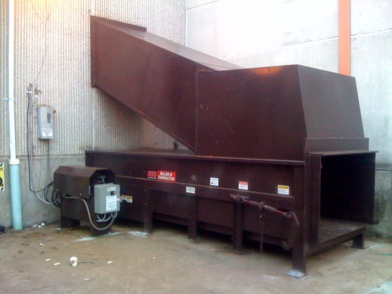 Large, dark brown industrial trash compactor in a concrete setting.