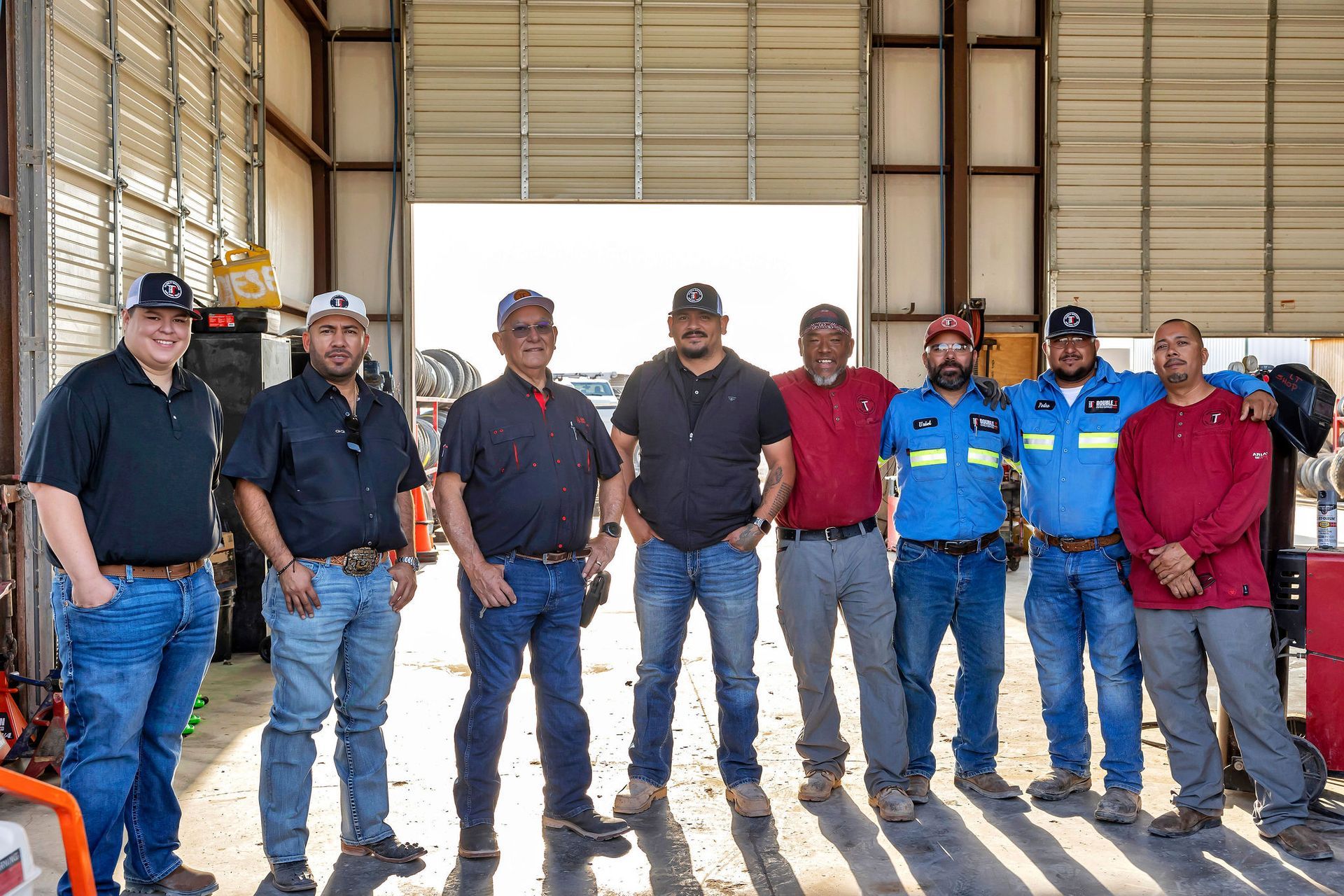 Group of nine men standing inside a garage. Some are wearing work uniforms. Sunlight streams in from the open doorway.