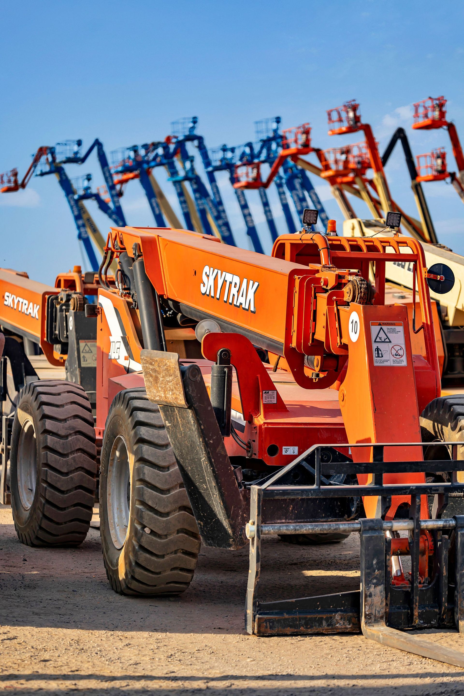 Orange SkyTrak forklift with black forks, in front of several blue boom lifts under a clear sky.