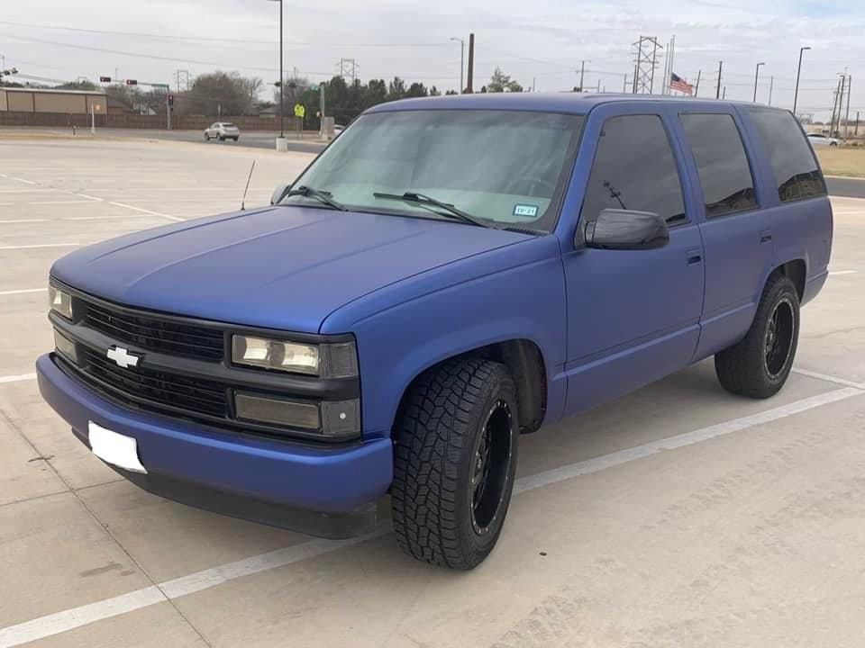Blue matte-finished Chevrolet Tahoe SUV with black wheels parked on a paved lot under an overcast sky.