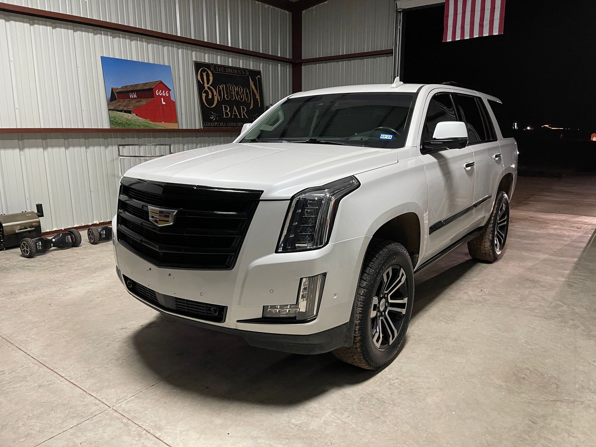 A white Cadillac Escalade SUV parked inside a garage with a red barn painting and American flag.