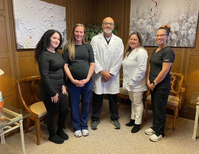 Five people pose together in an office setting. They are smiling, and two women are pregnant.