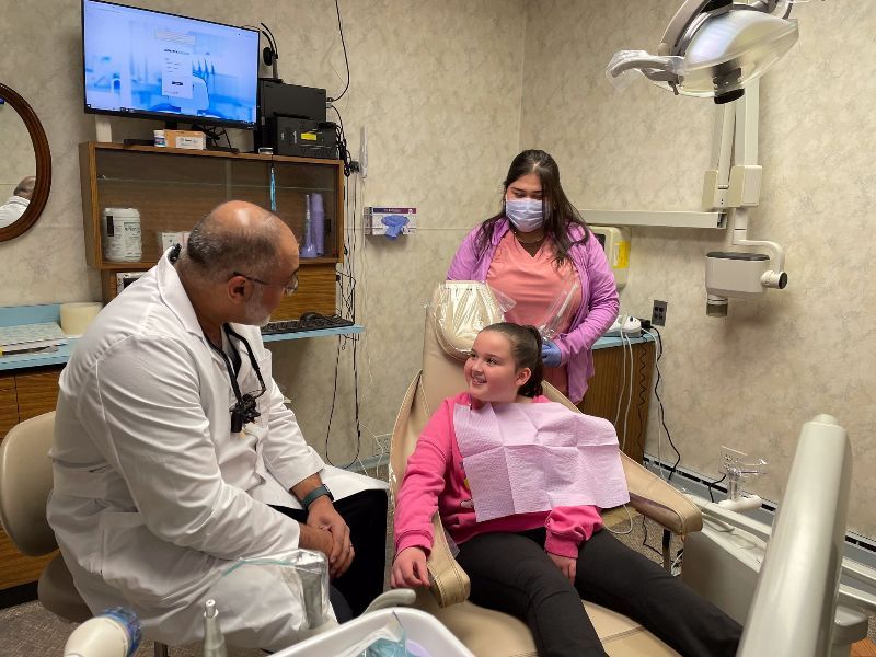 Dentista, asistente y jovencita en la consulta. La joven sonríe en la silla; el dentista habla; el asistente está detrás de ella.