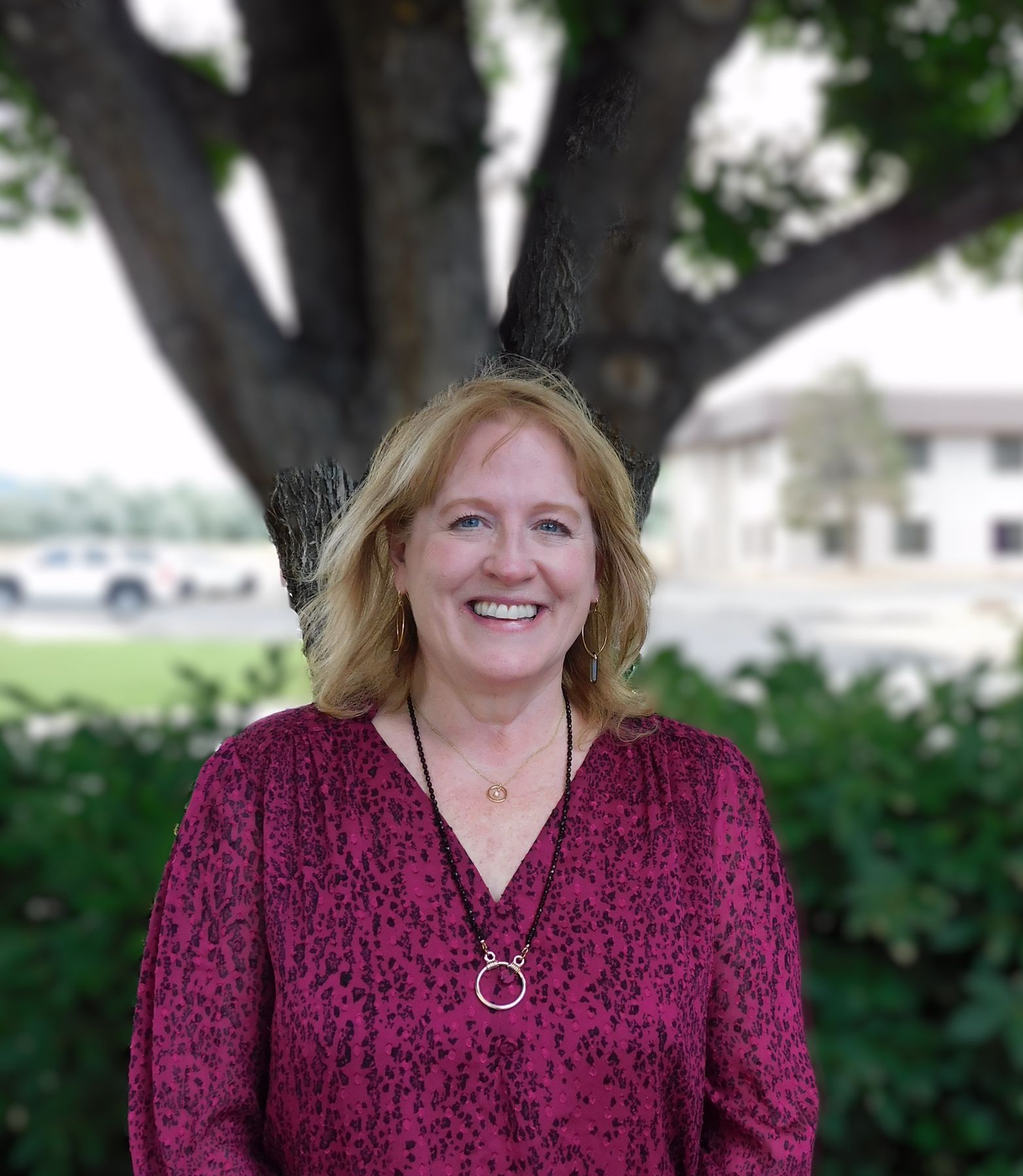 A woman in a red shirt and necklace is smiling in front of a tree.