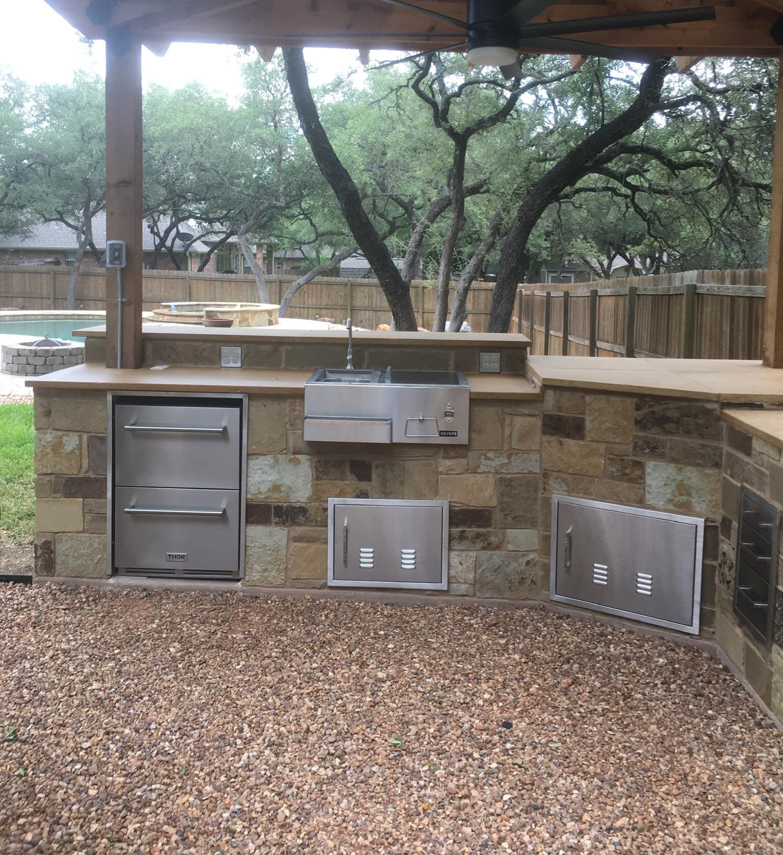 Outdoor kitchen with stone counters, stainless steel appliances, and pebble flooring.