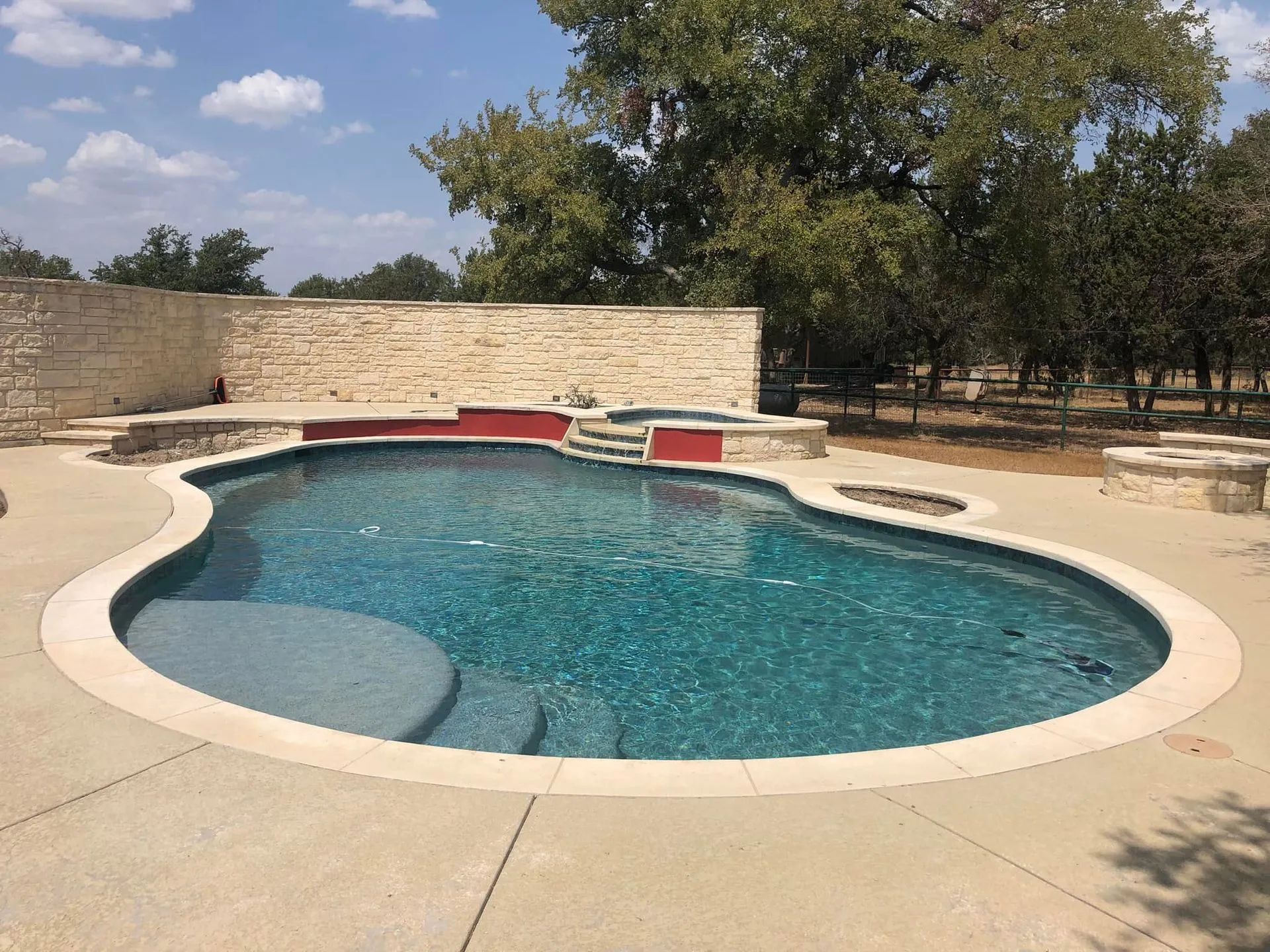 Empty backyard swimming pool with blue water, beige stone deck, and trees in the background
