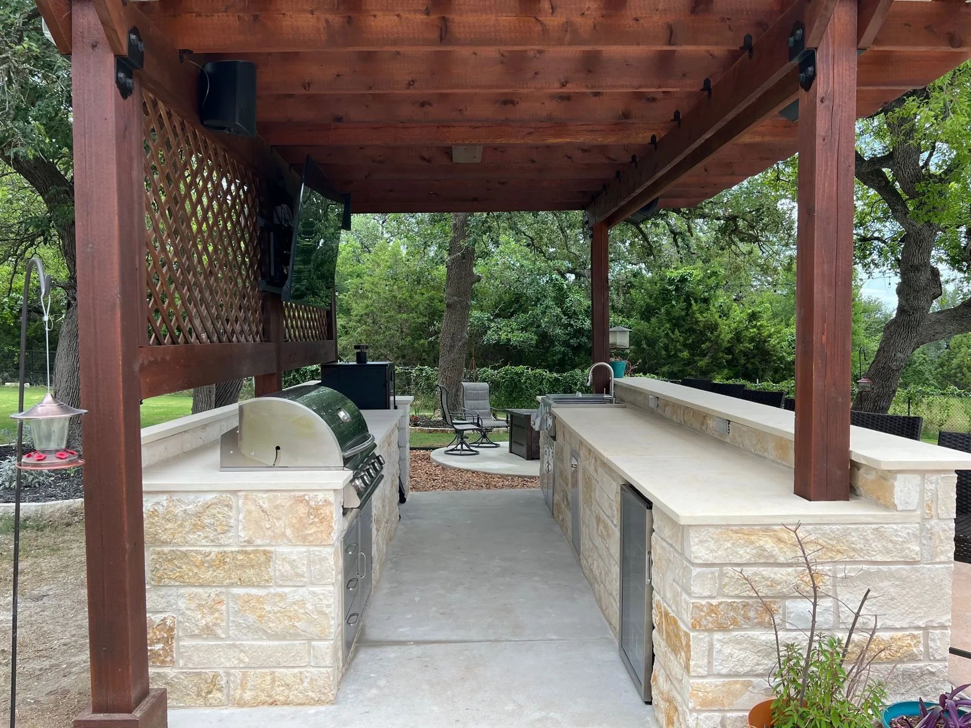 Covered outdoor grilling area with stone counters, built-in grill, and wooden pergola surrounded by trees.