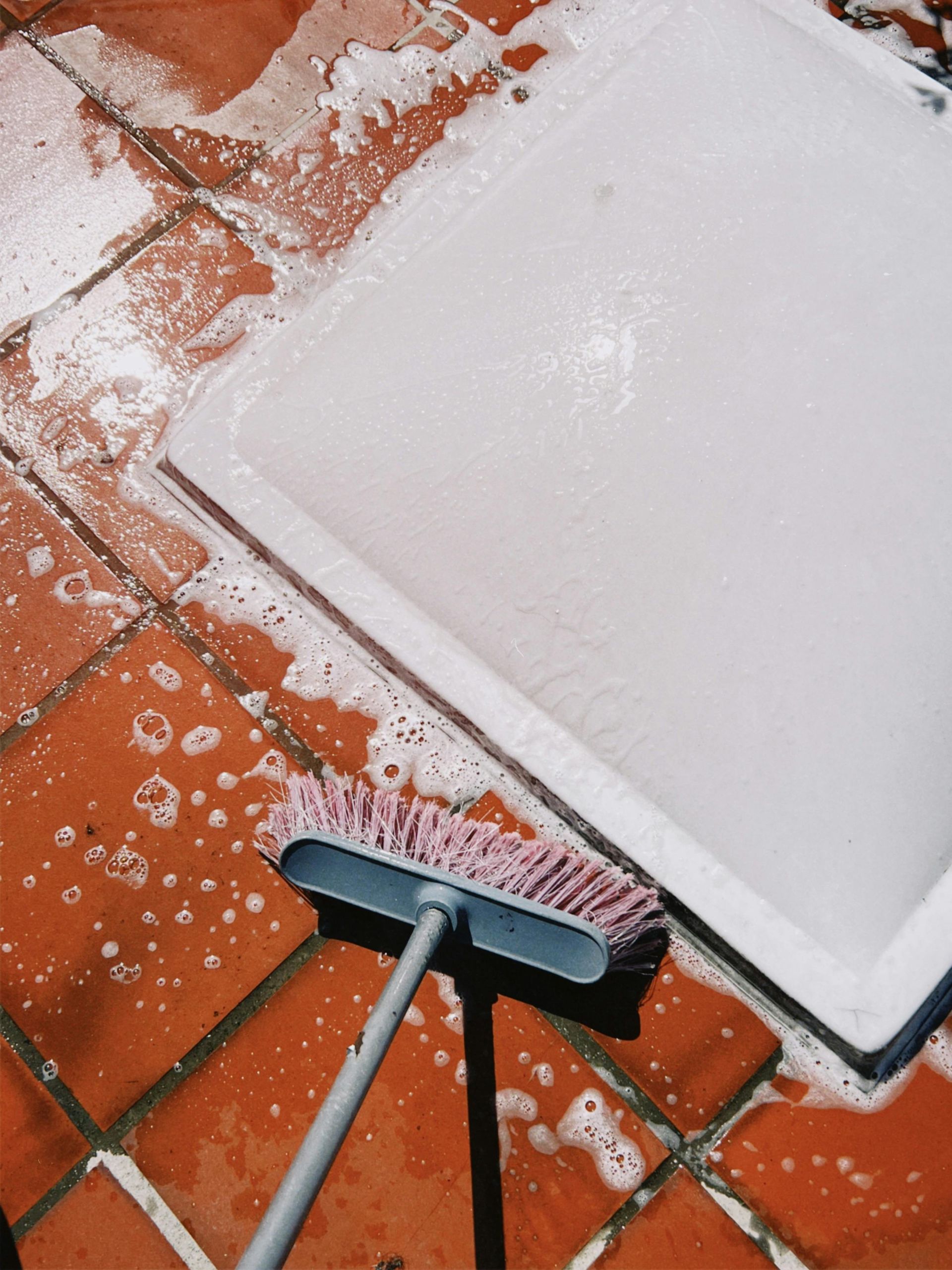 A person cleaning a white surface with soapy water, a broom on red tiled floor.