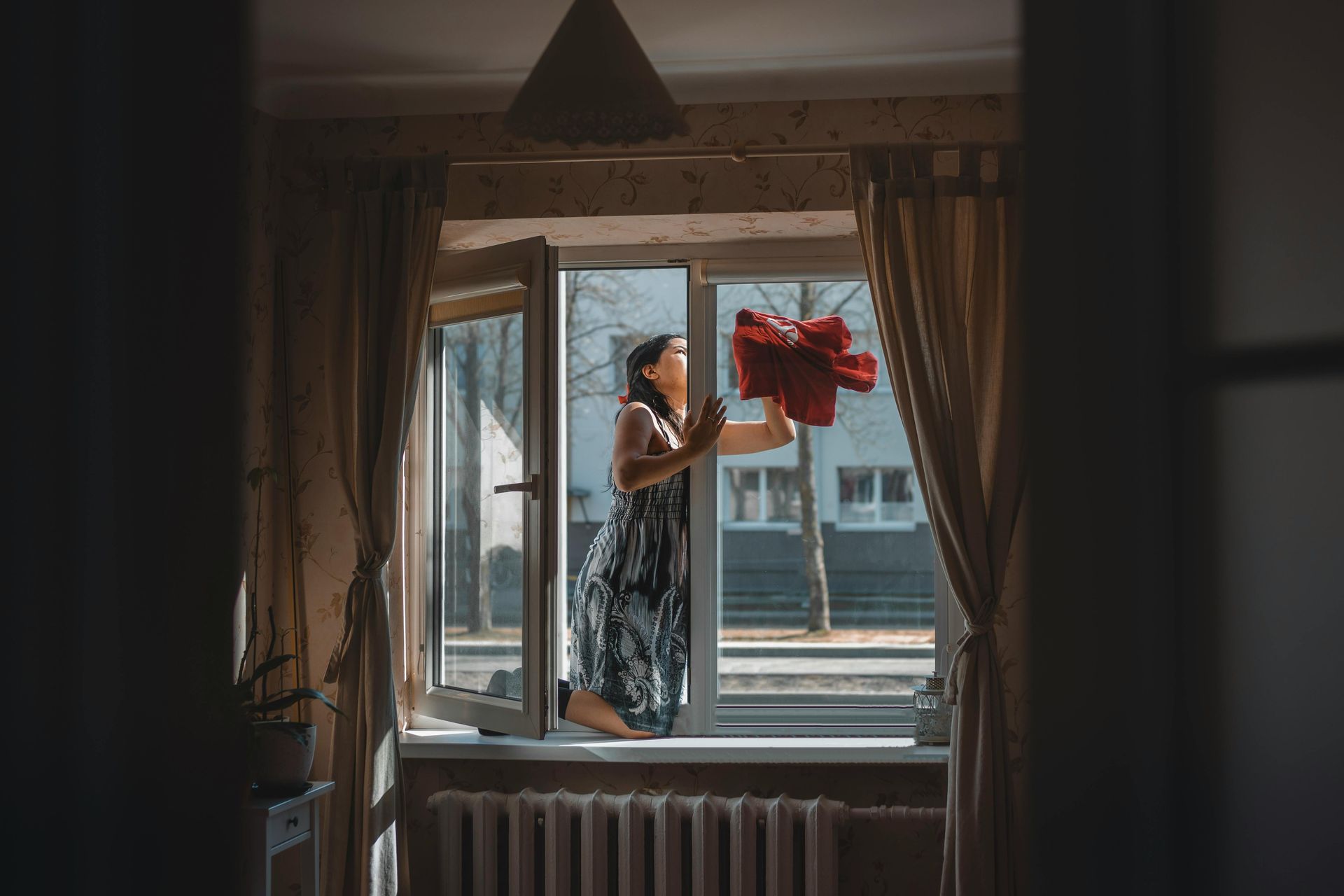 Woman in floral dress leaning out open window, holding red flowers. Sunlight, building exterior.