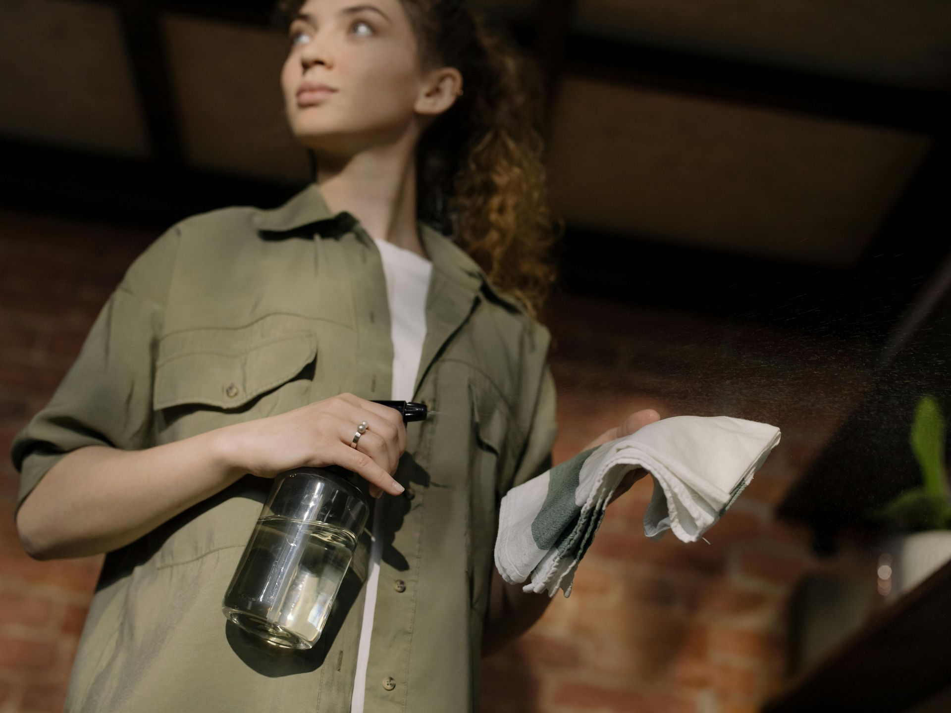 Woman spraying cleaner, wiping with a cloth. Brick wall background.