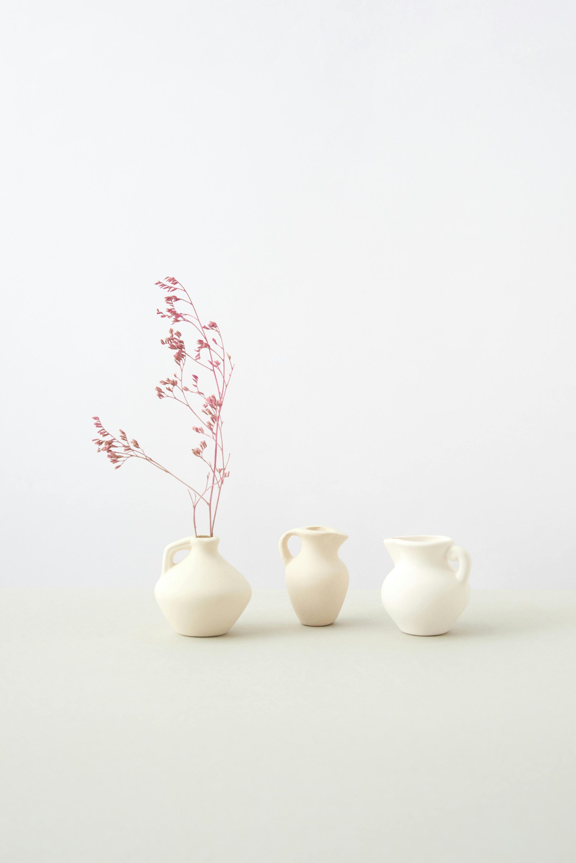 Three white ceramic vases, with a sprig of reddish dried flowers in one.