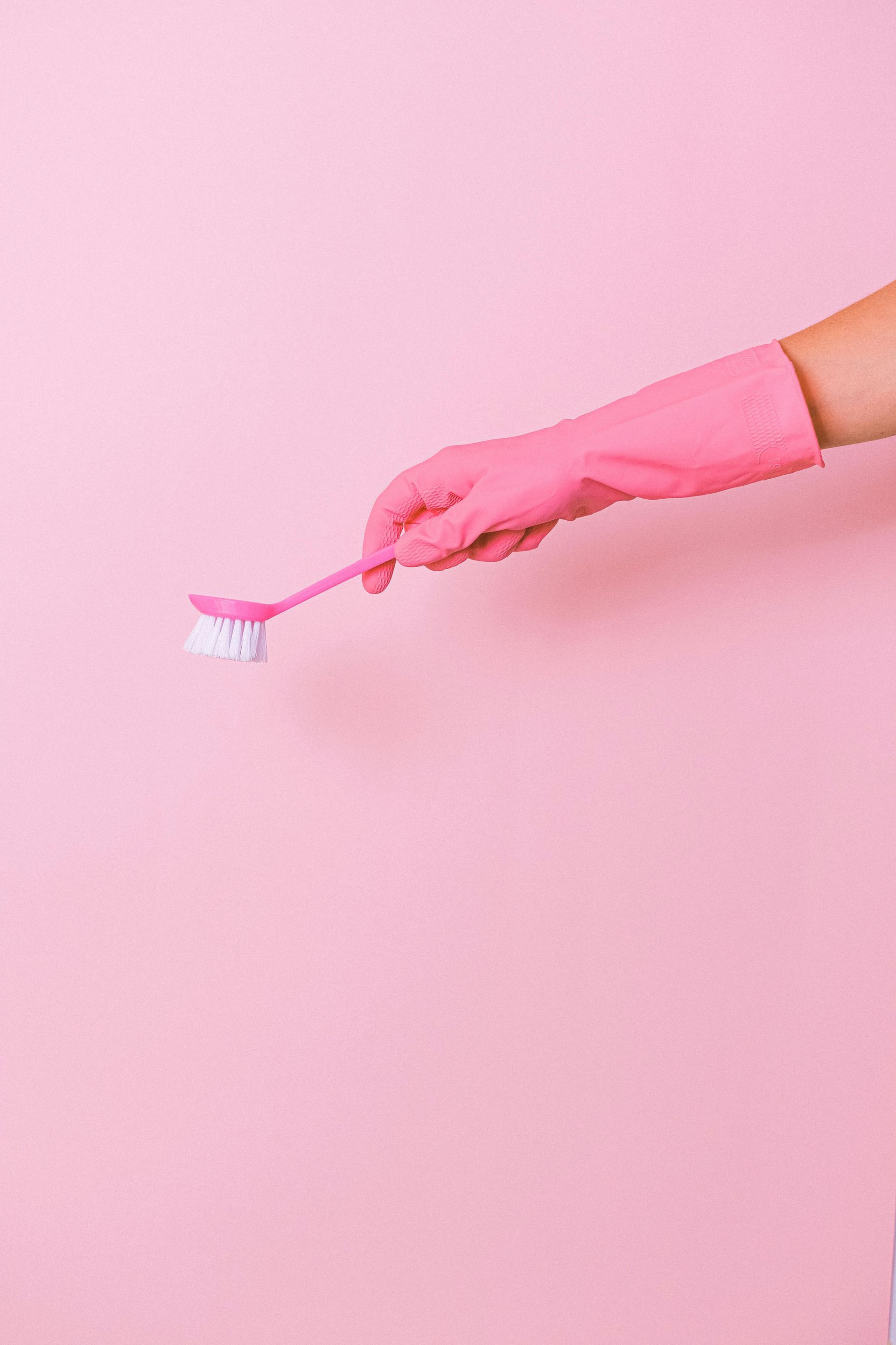 Pink-gloved hand holding a pink scrub brush against a pink background, cleaning.